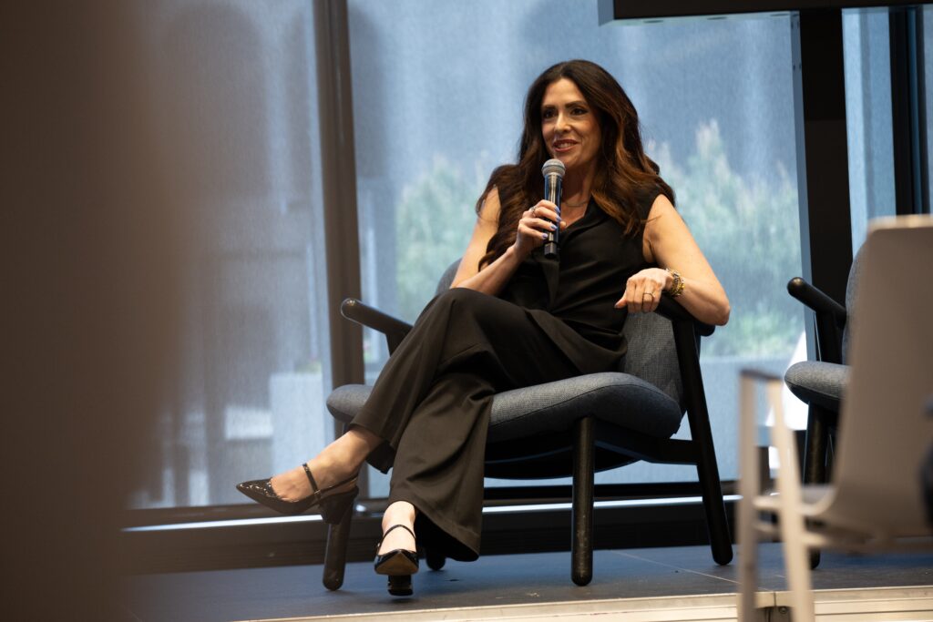 A woman dressed in black sits on a chair, holding a microphone and speaking about actionable data and AI, with large windows and natural light in the background.