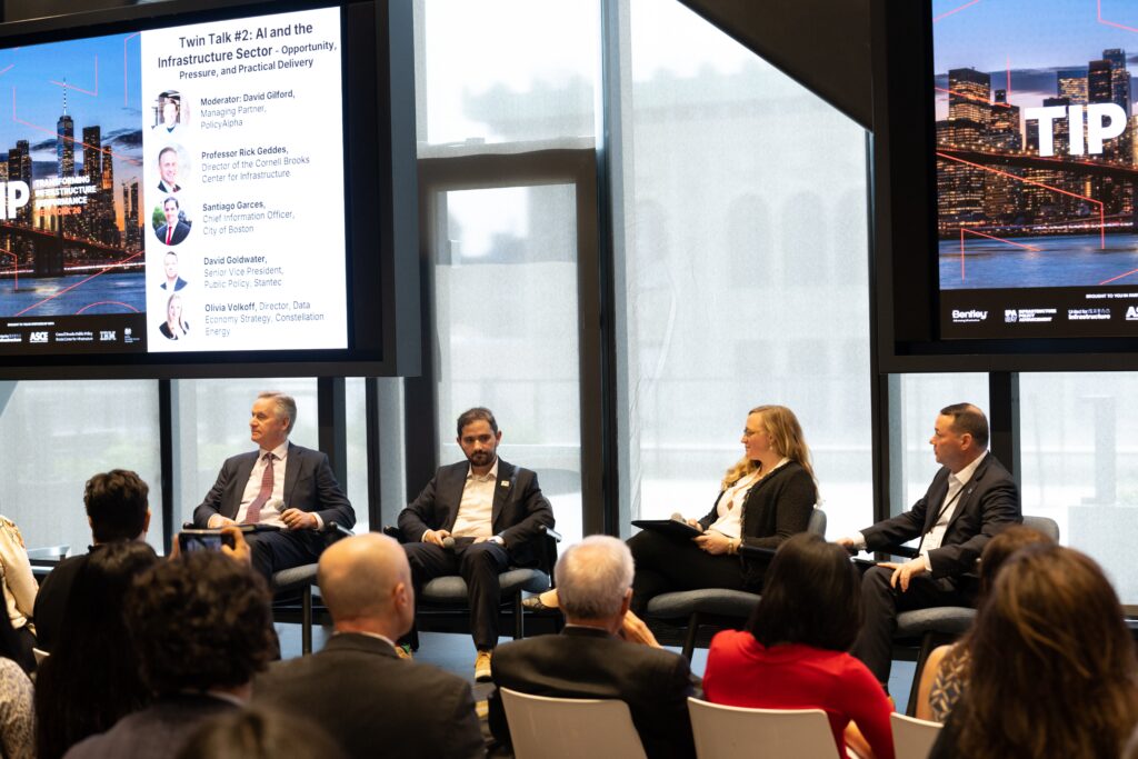 Four panelists sit onstage discussing AI and actionable data in the U.S. infrastructure sector at a conference, with large screens displaying event information behind them.