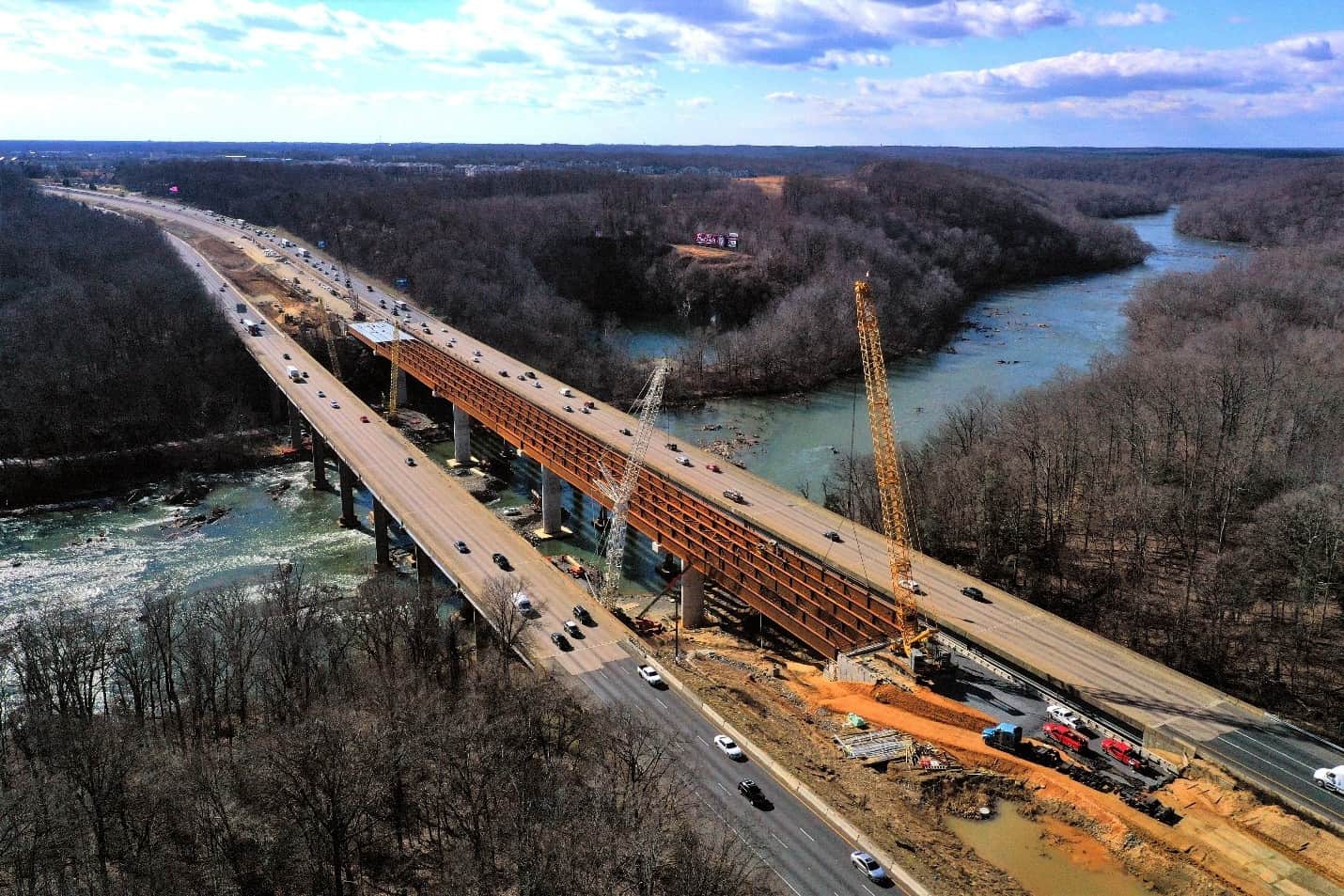 Aerial view of a highway bridge under construction over a river, showcasing cranes, construction vehicles, and ongoing traffic on adjacent lanes&mdash;highlighting modern design and innovative construction techniques.