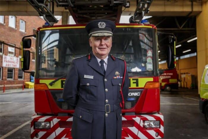 Fire chief in uniform stands in front of a fire truck parked inside a fire station.