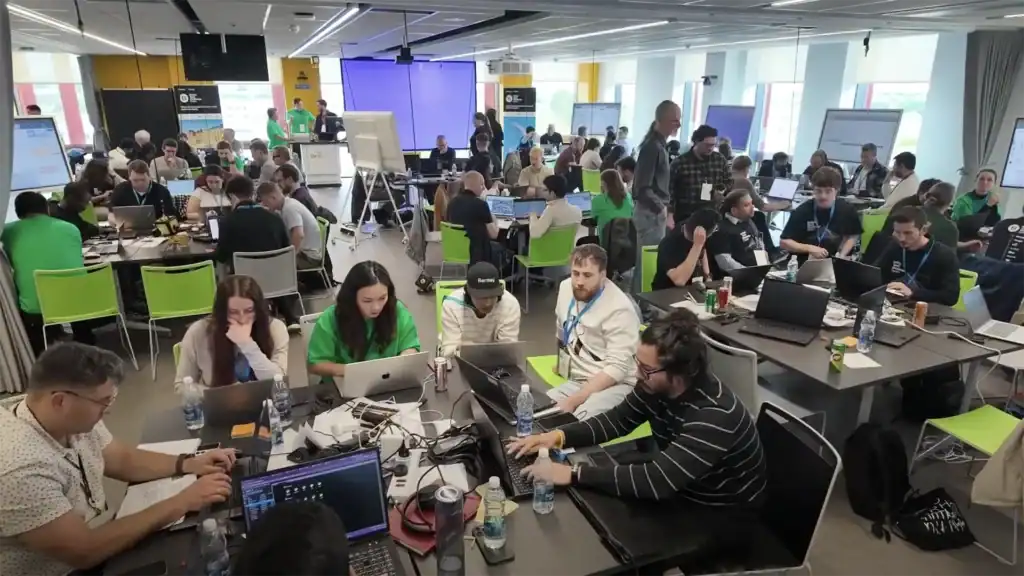 A large group of people work on laptops and collaborate in a busy, modern conference room with bright lighting and open seating.