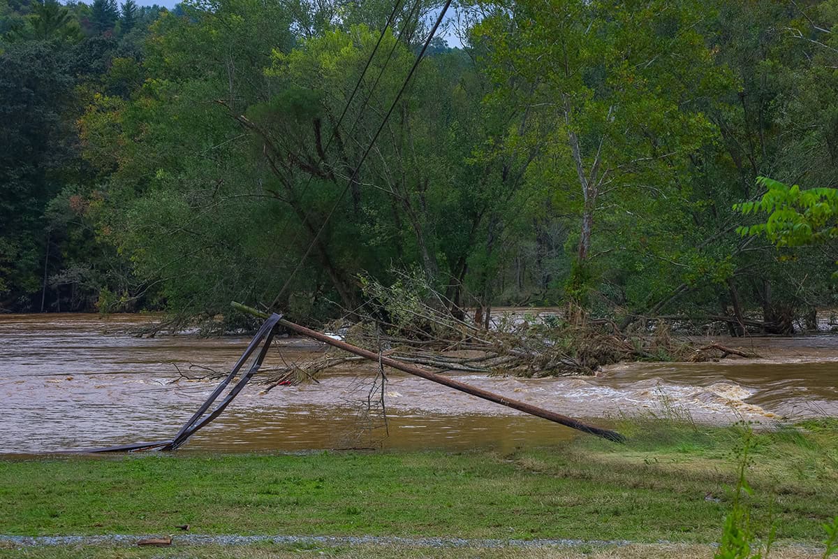 A flooded river with muddy water flows through trees; a broken utility pole and sagging power lines lean across the riverbank, presenting visualization challenges for iTwin Engage.