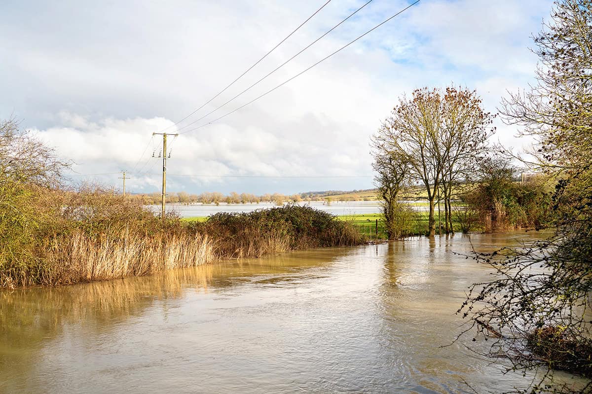 Flooded rural landscape with water covering a road, partially submerged bushes and trees, and power lines overhead under a cloudy sky&mdash;highlighting the visualization challenges that iTwin Engage helps address.