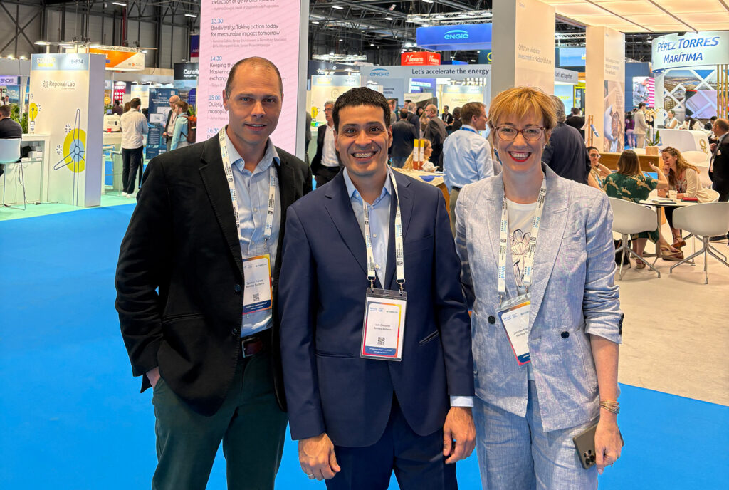 Three professionals posing for a photo at a trade show on a blue exhibition floor, badges visible around their necks.