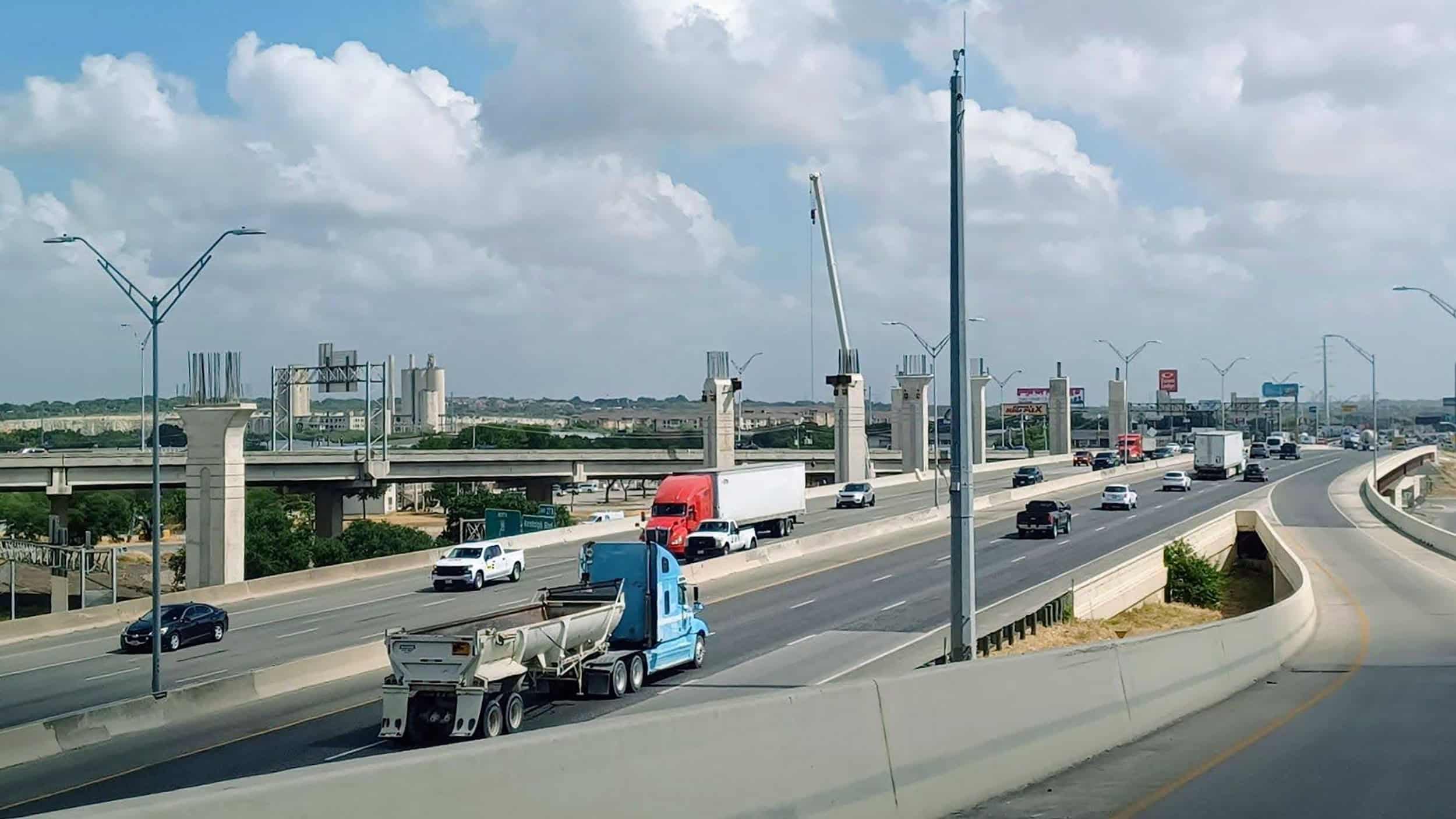 Cars and trucks travel on a multi-lane highway with an overpass under construction in the background under a partly cloudy sky.