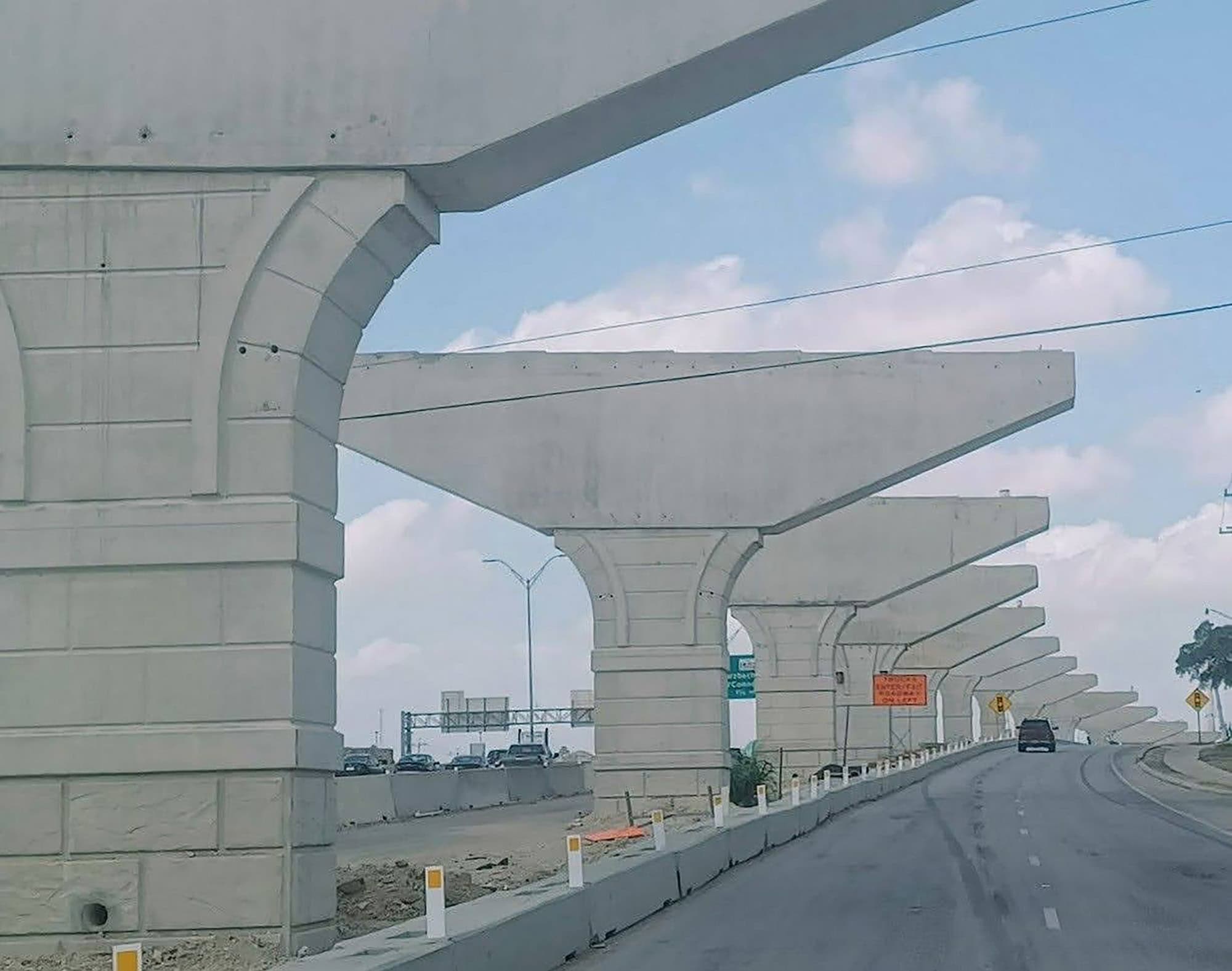 Unfinished elevated highway supports stand alongside a road under construction, with cars and road signs visible in the background under a partly cloudy sky.
