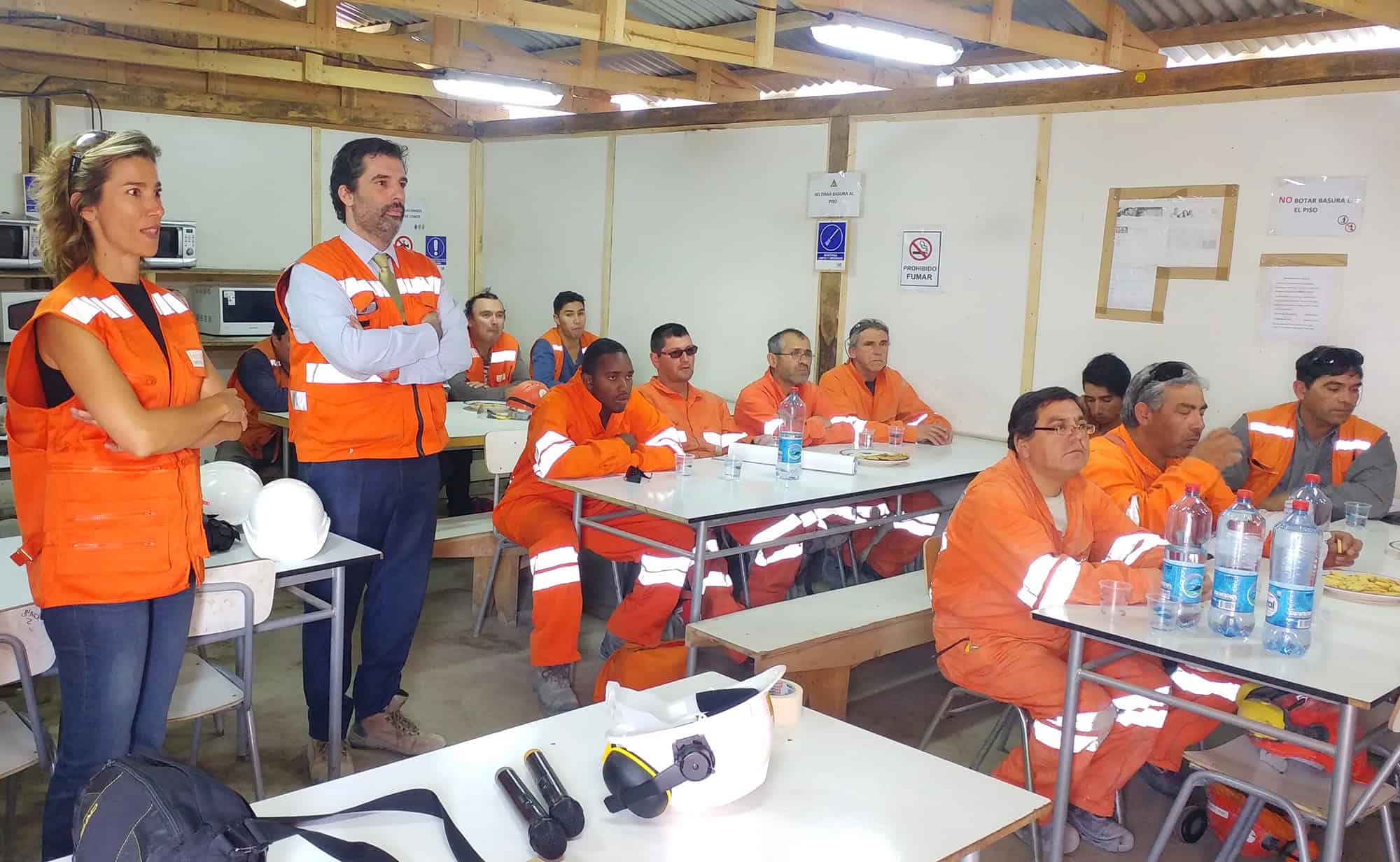 Workers in orange vests sit and listen during a meeting inside a break room with wooden benches and tables, hard hats, and water bottles visible.