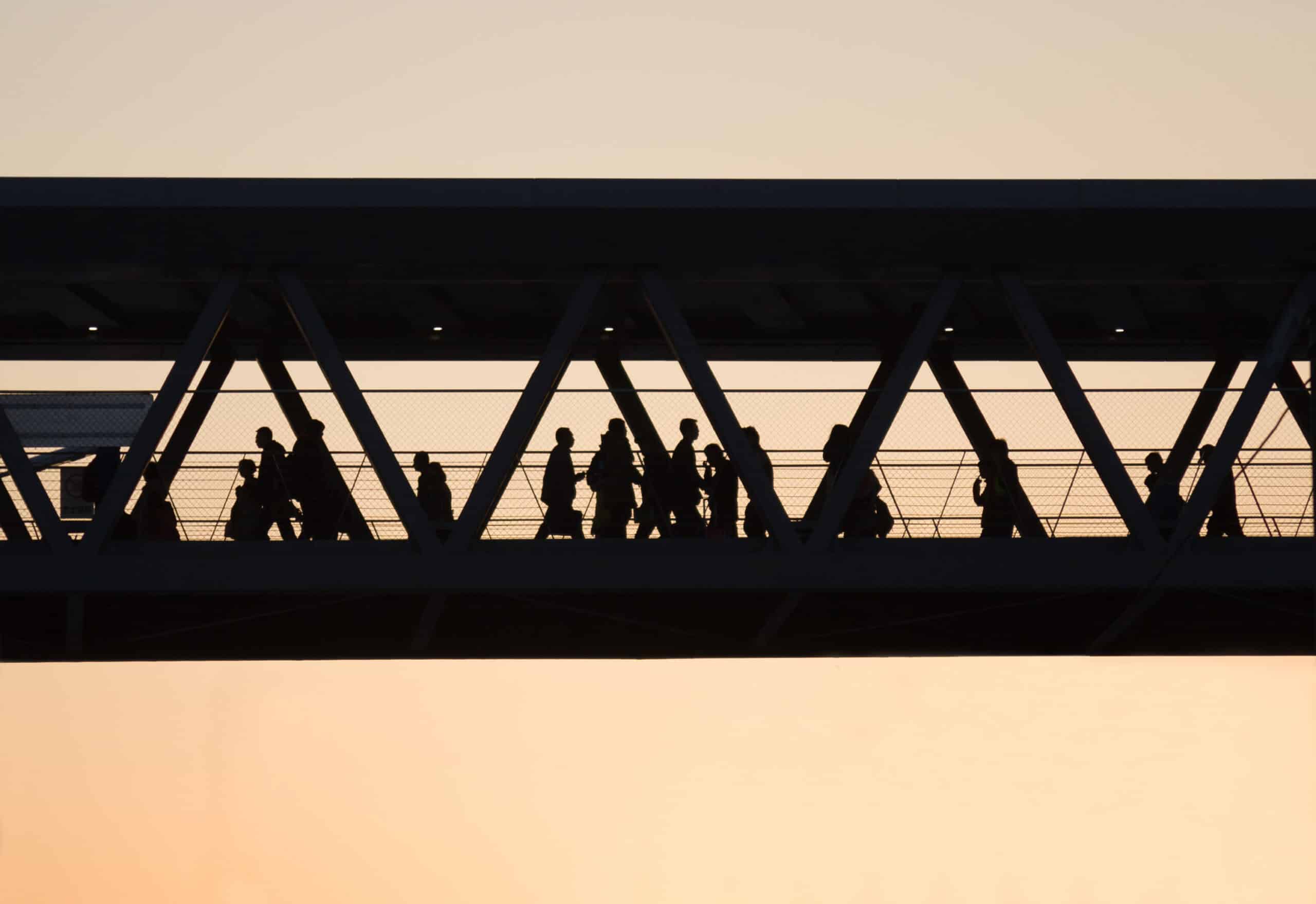 Silhouettes of people walking across an enclosed pedestrian bridge at sunset evoke the quiet transformation brought by AI disruption and technology infrastructure shaping modern city life.