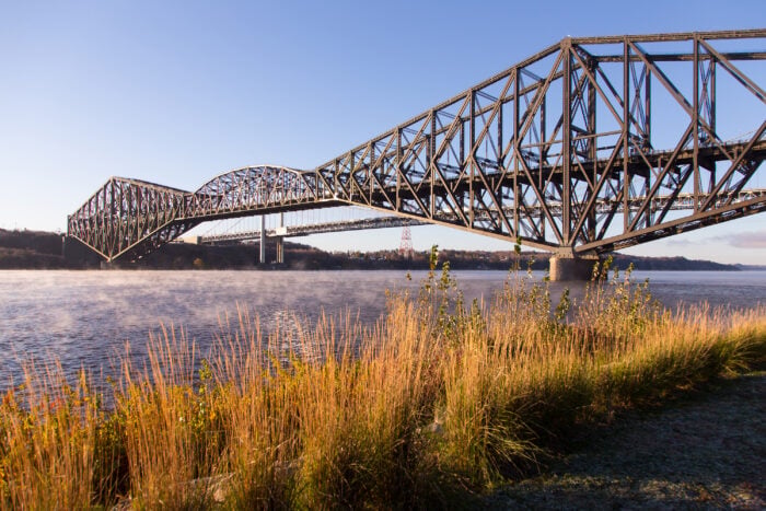 A large steel cantilever bridge spans over a wide river, with tall grass in the foreground and a clear blue sky overhead.