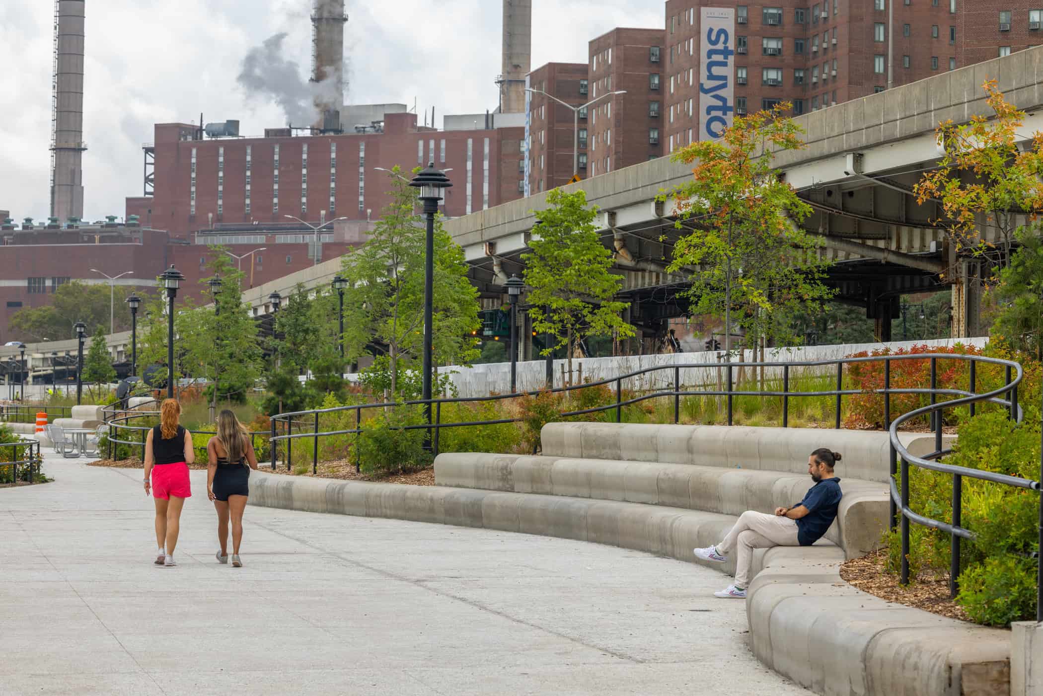 Two women walk along a curved pedestrian path while a man sits on tiered concrete seating; buildings and smokestacks are visible in the background.
