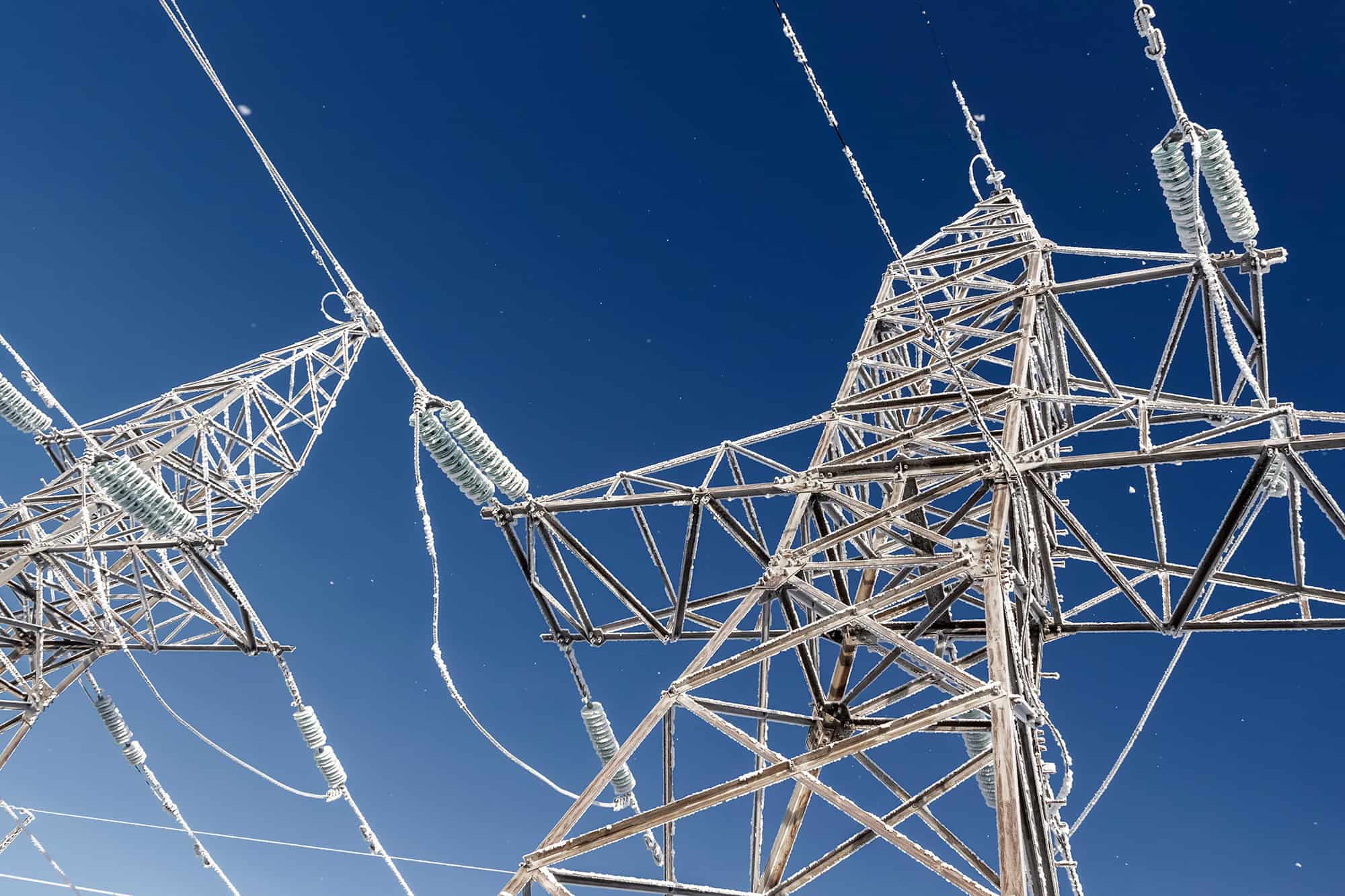 Two metal electricity pylons covered in frost, with wires extending across a clear blue sky.