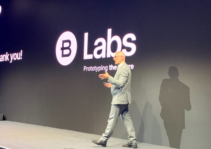A man in a light gray suit walks on stage in front of a black wall displaying the words Labs Prototyping the Future and a logo with a B inside a circle.