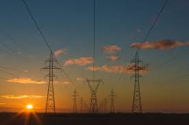 High-voltage power lines and electrical towers stretch across a landscape at sunset, with a clear sky and scattered clouds.