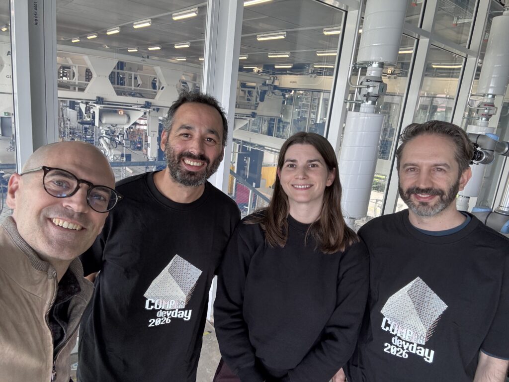 Four people standing indoors in front of industrial equipment, three wearing matching CONRAD devday 2026 shirts, smiling at the camera during the Swiss Symposium focused on design and build innovations.