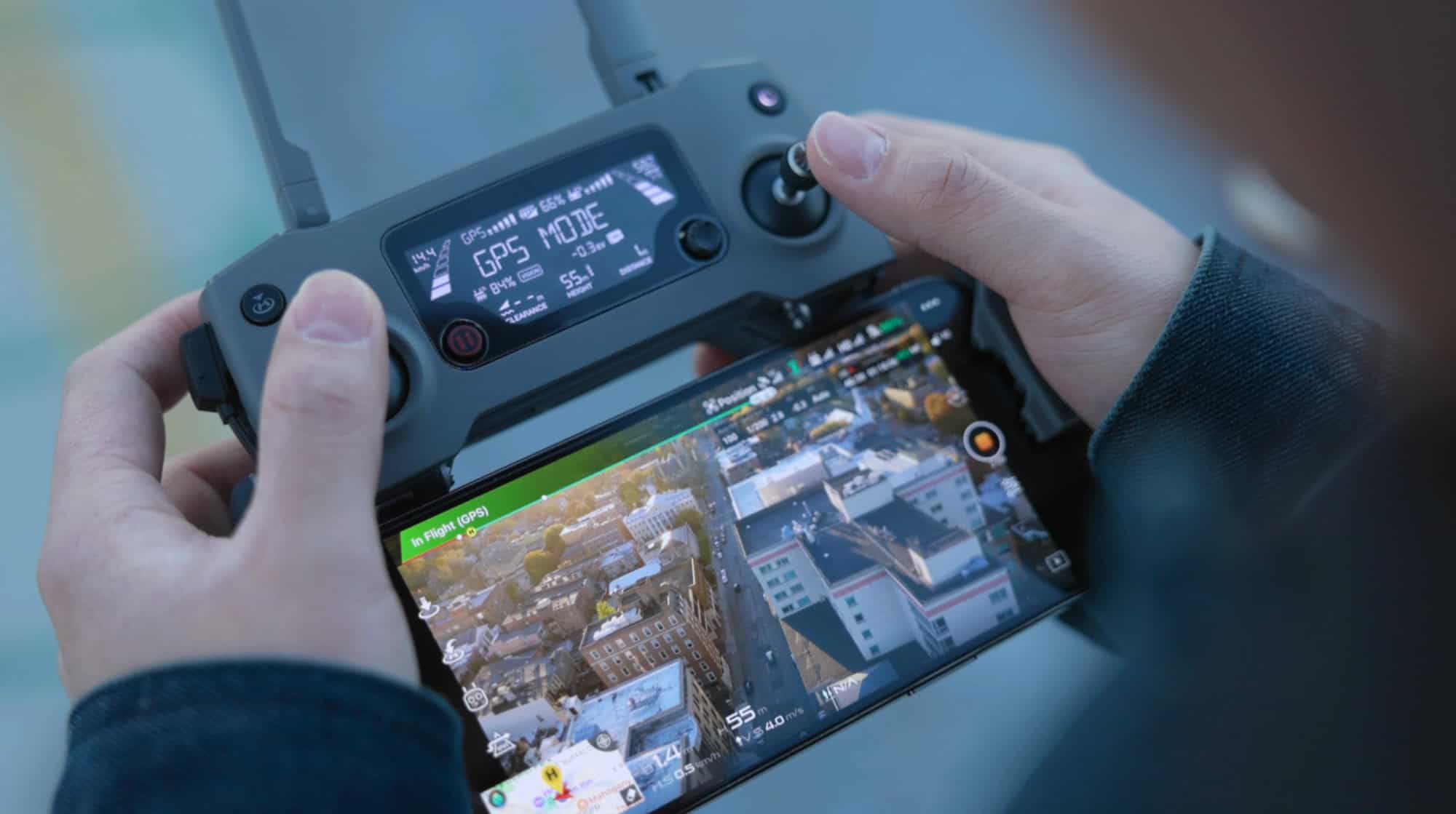 Close-up of hands operating a drone remote controller with a smartphone display showing aerial view of a cityscape.