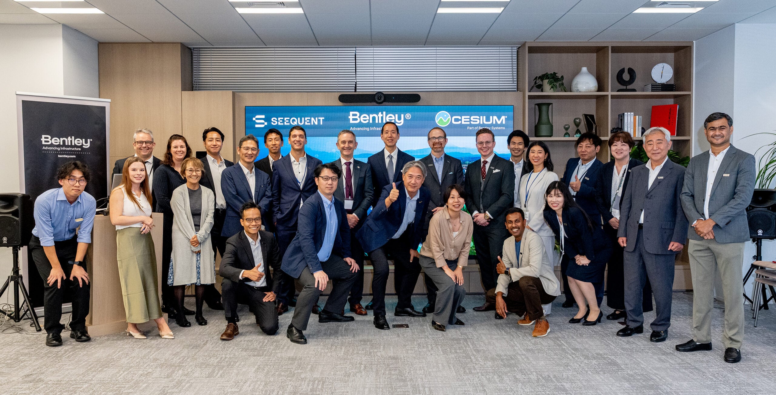 A group of business professionals poses together in an office space, with some people smiling and making celebratory gestures in front of a display with company logos.