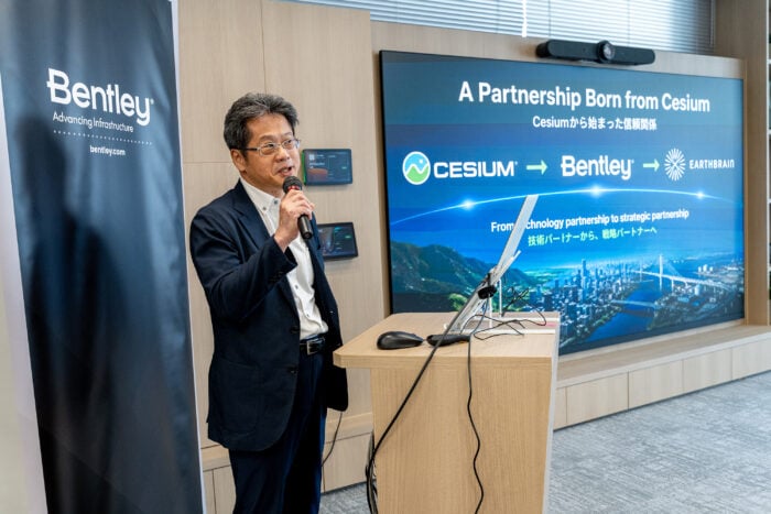 A man in a suit speaks at a podium with a microphone. Behind him, a screen displays a partnership announcement between Cesium, Bentley, and EarthBrain.