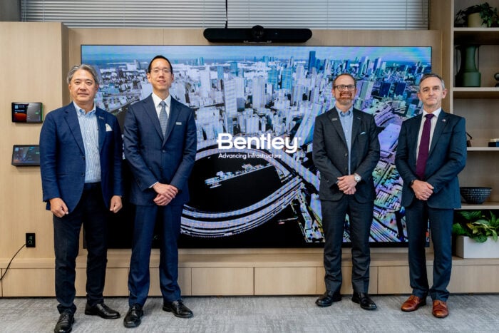 Four professionals in suits posing in front of a large Bentley banner in a corporate office environment.