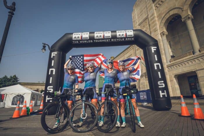 Four cyclists in matching gear celebrate at the race finish line, holding American and British flags, with their bicycles in front. A banner above reads "The World's Toughest Bicycle Race."