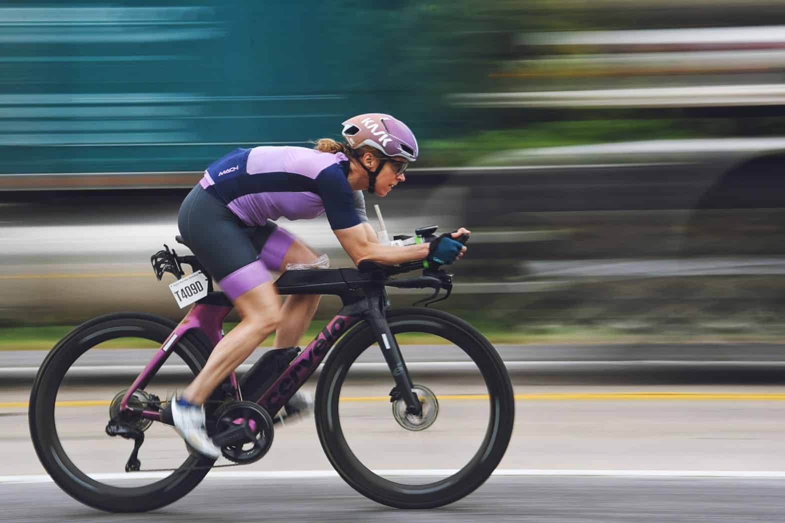 Person in purple cycling attire and helmet rides a black bicycle on a road, with a blurred background indicating high speed.