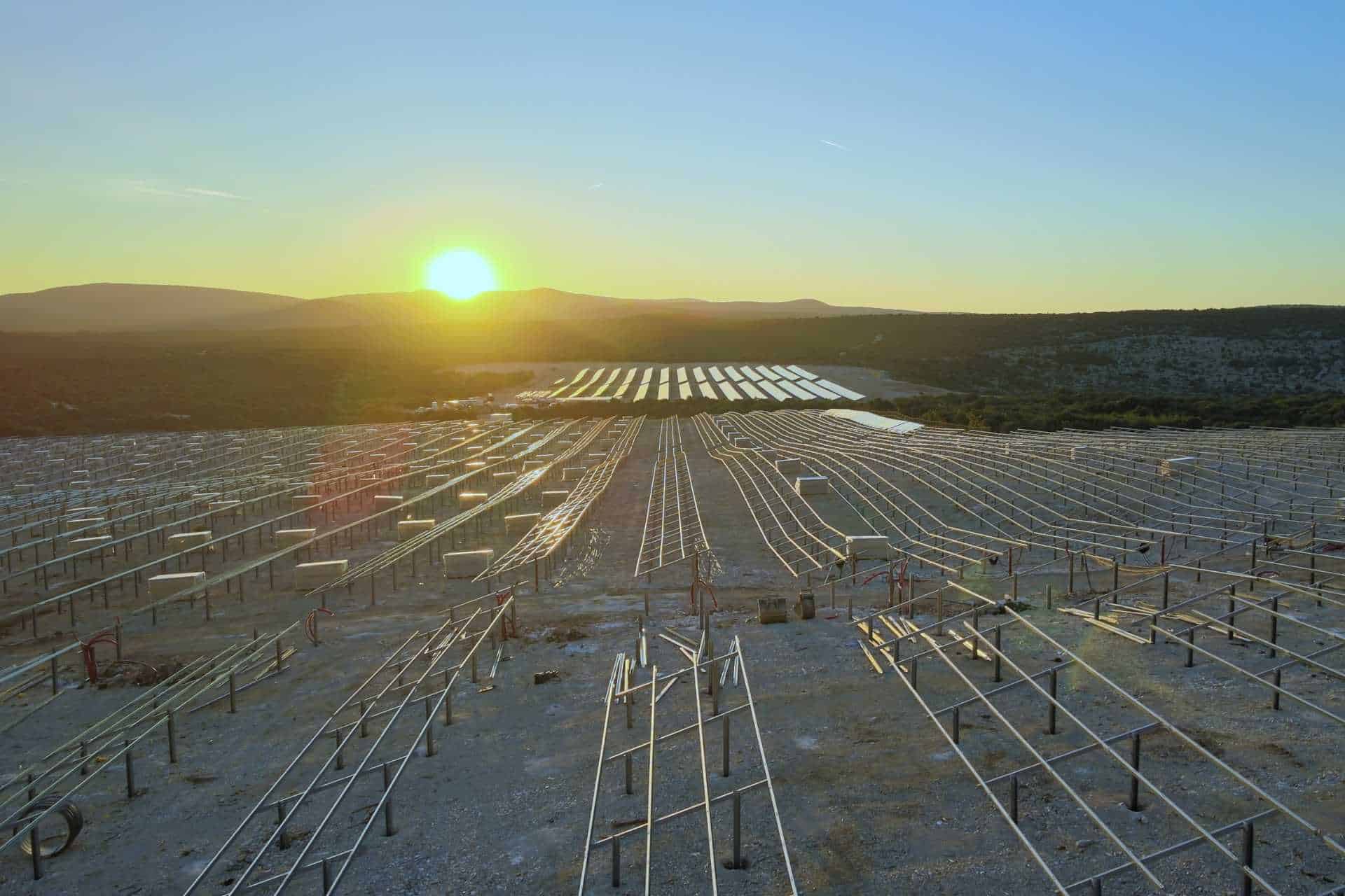 Metal frames for solar panels are arranged in rows on a large outdoor site at sunset, with mountains visible in the background.