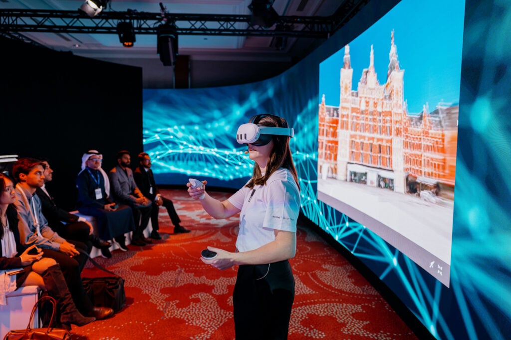 Liana O’Cleirigh wearing a VR headset stands and interacts with a controller in front of an audience, with a large screen displaying a digital image of a building in the background.