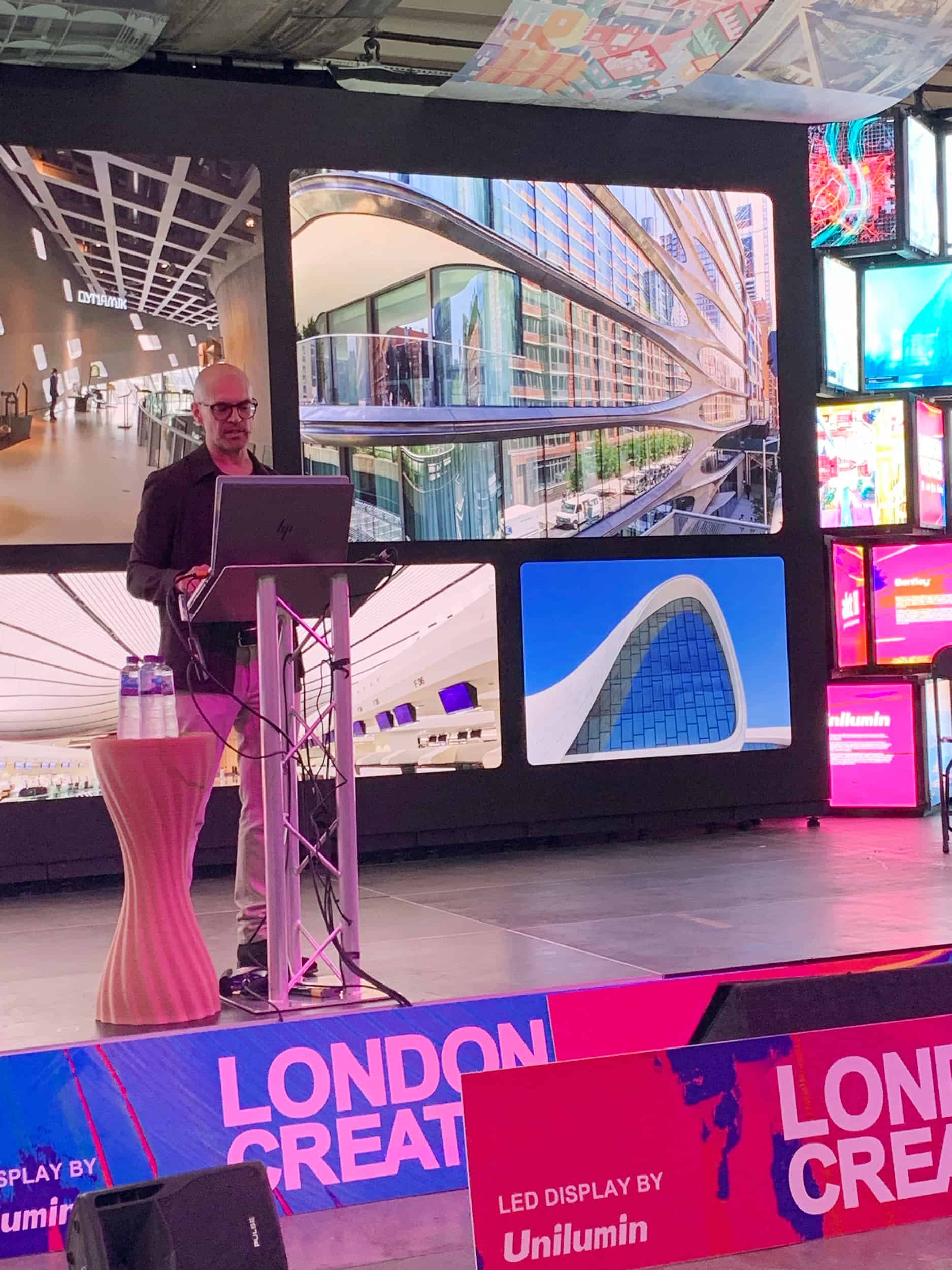 A person stands at a podium giving a presentation on architecture, with large modern building images displayed on screens behind.