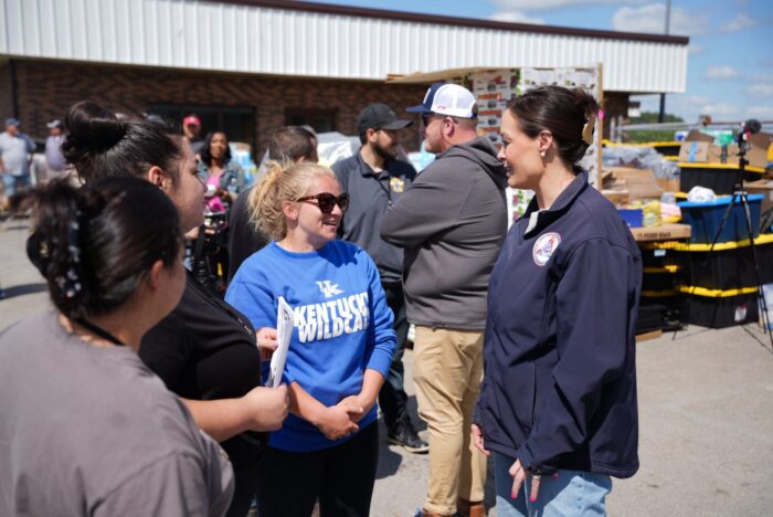 Kentucky Lt. Gov. Jacqueline Coleman and a group of volunteers stand and talk outside a distribution center, with supplies and boxes visible in the background on a sunny day.