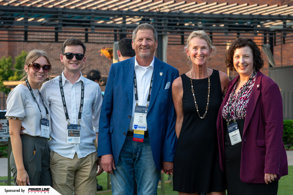 Five people stand outdoors at a tech event, wearing business casual attire and name badges, smiling at the camera.