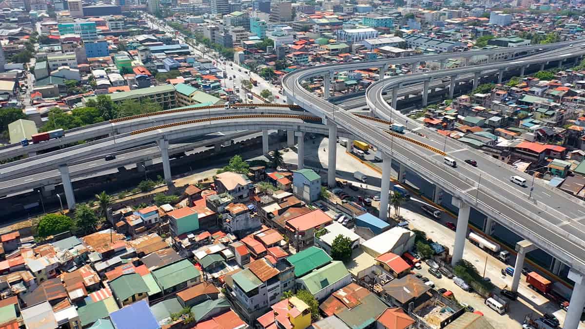 Aerial view of a multi-level highway interchange, part of tomorrow's roads, above a densely populated urban area with colorful rooftops and various vehicles navigating the digital infrastructure below.
