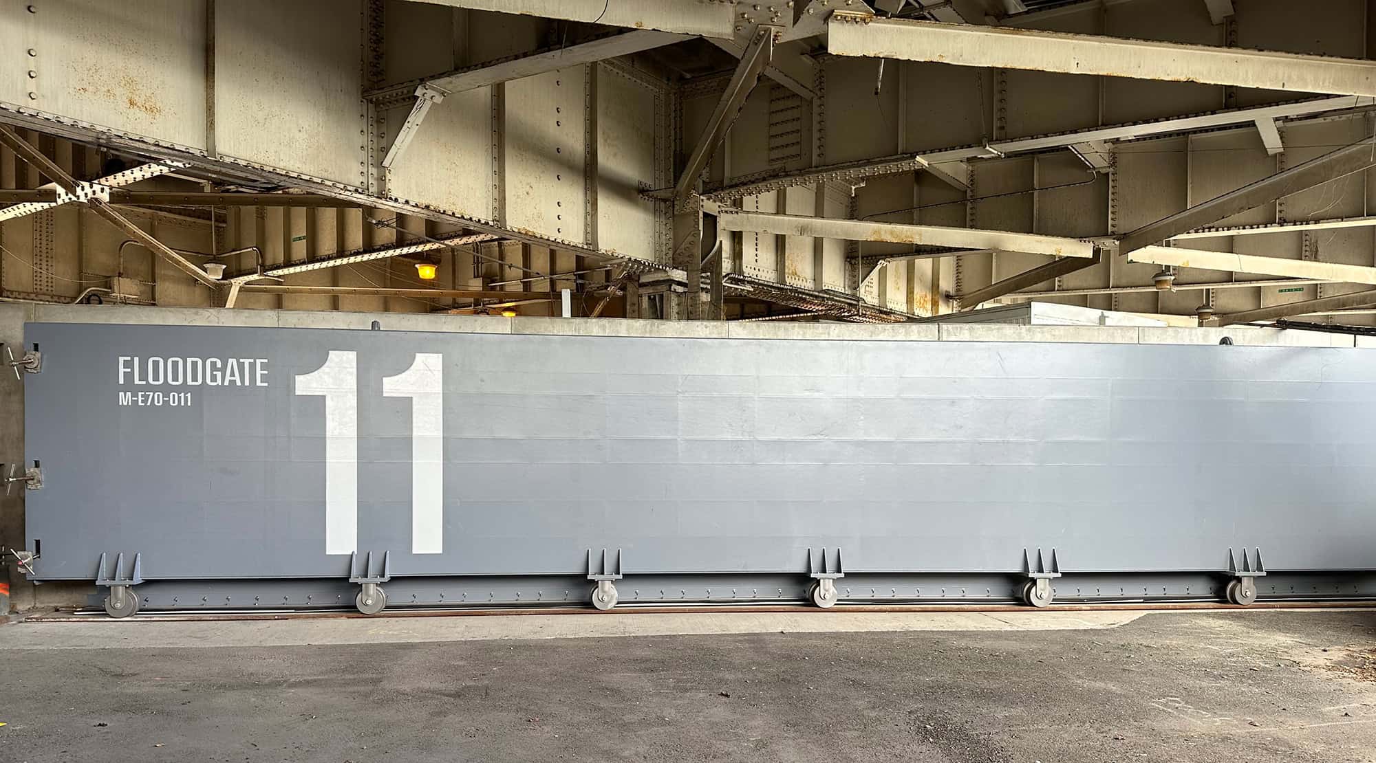 A large, gray floodgate labeled FLOODGATE MF01-011 and the number 11 stands beneath a metal bridge structure.