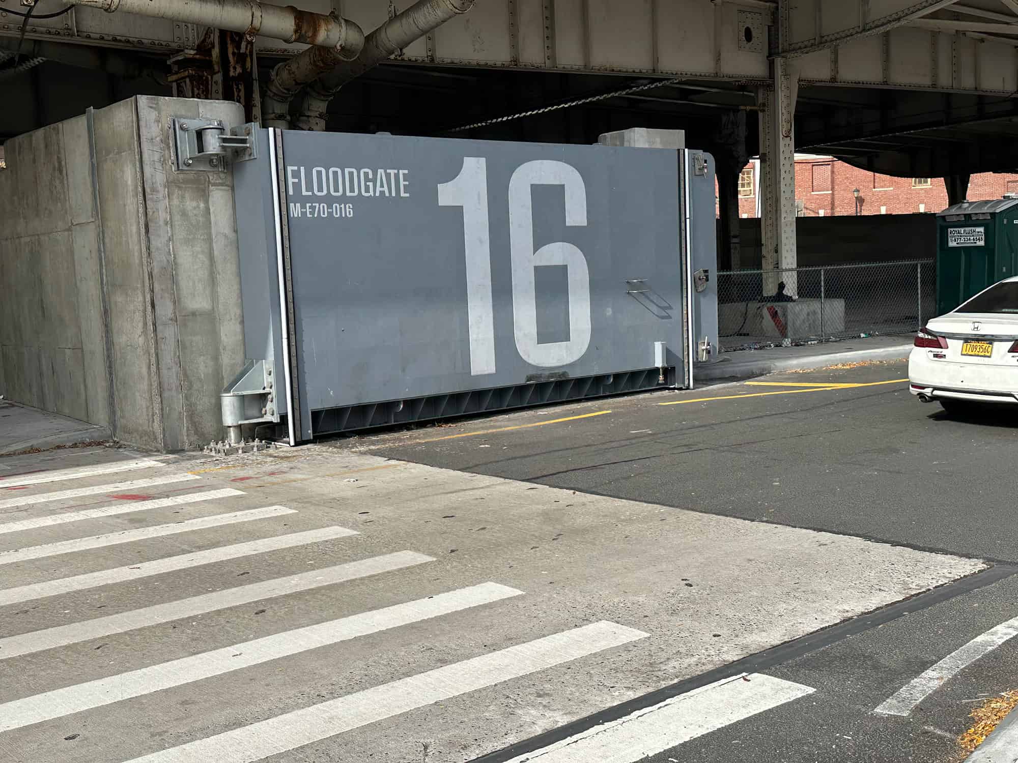 Large gray floodgate labeled FLOODGATE MF-070-016 16 is installed under an overpass near a crosswalk and parked car.