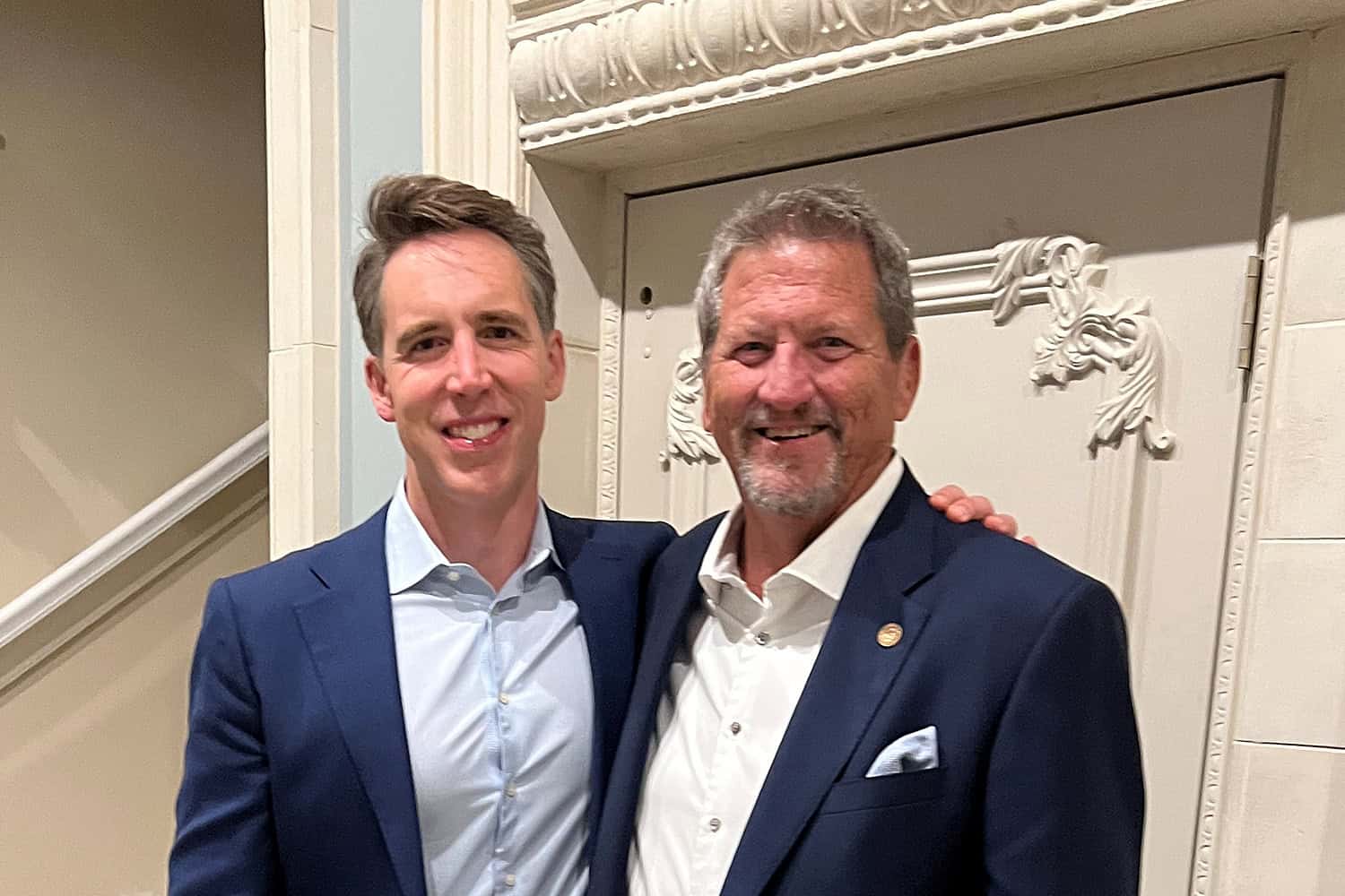 Two men in suits stand side by side, smiling at the camera in front of an ornate white door.