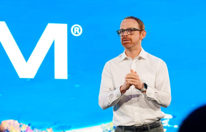 A person in a white shirt and glasses speaks on stage with a blue background and a partial letter M visible.