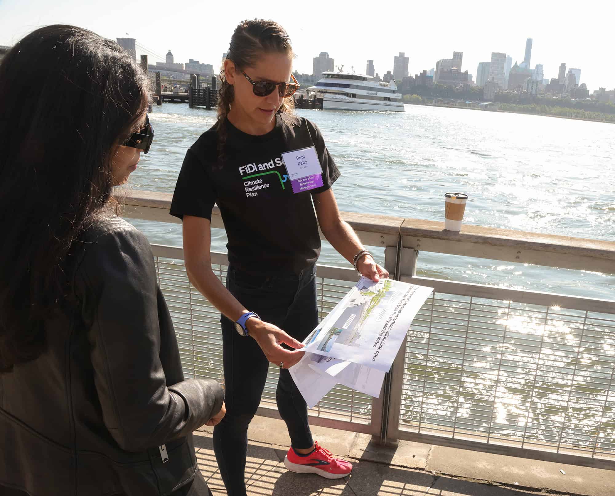 Two women stand by a waterfront railing; one holds and examines a map or plan while the other listens. A ferry and city skyline are visible in the background.