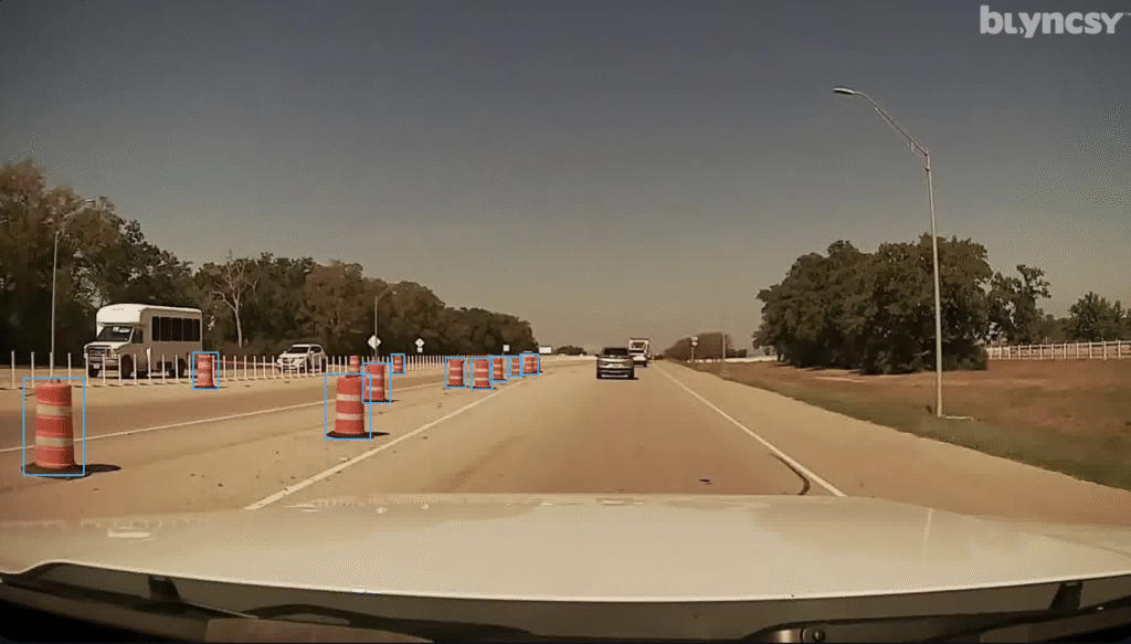 A view from a vehicle driving on a divided highway showcases Alabama road infrastructure with orange construction barrels separating lanes and some vehicles ahead under a clear sky.