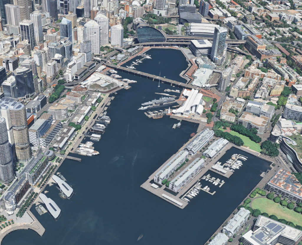 Aerial view of a harbor city waterfront featuring docks, boats, marinas, high-rise buildings, green spaces, and urban areas.