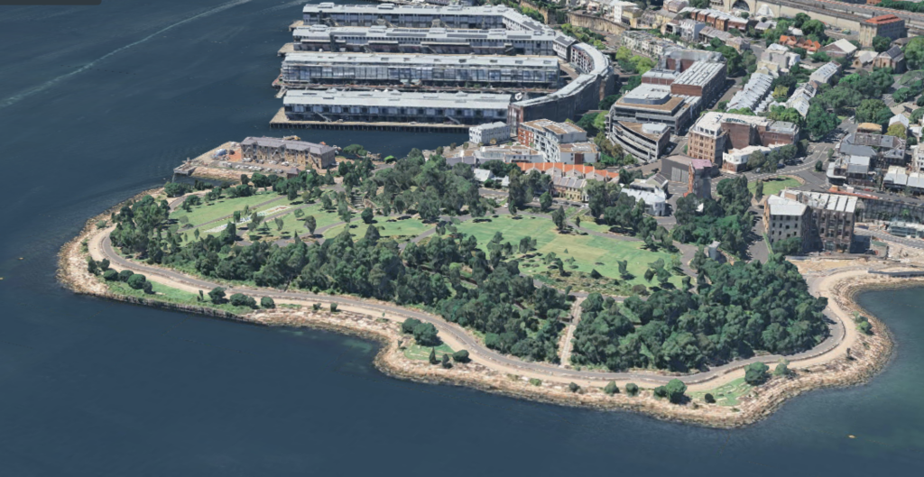 Aerial view of a green park with trees and paths on a small peninsula, surrounded by water and adjacent to a marina with docked buildings.