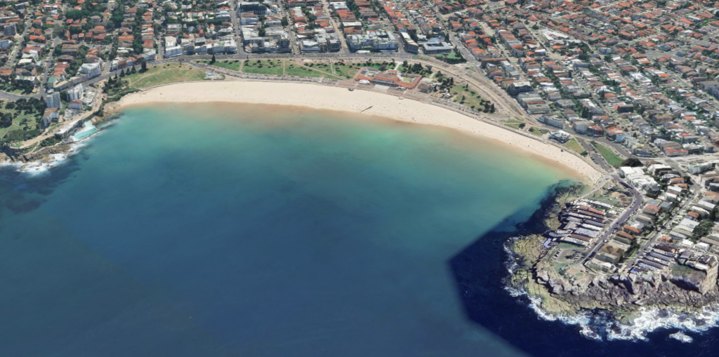 Aerial view of a sandy beach bordered by urban buildings, a coastal park, and clear blue-green water in the vibrant harbor city.