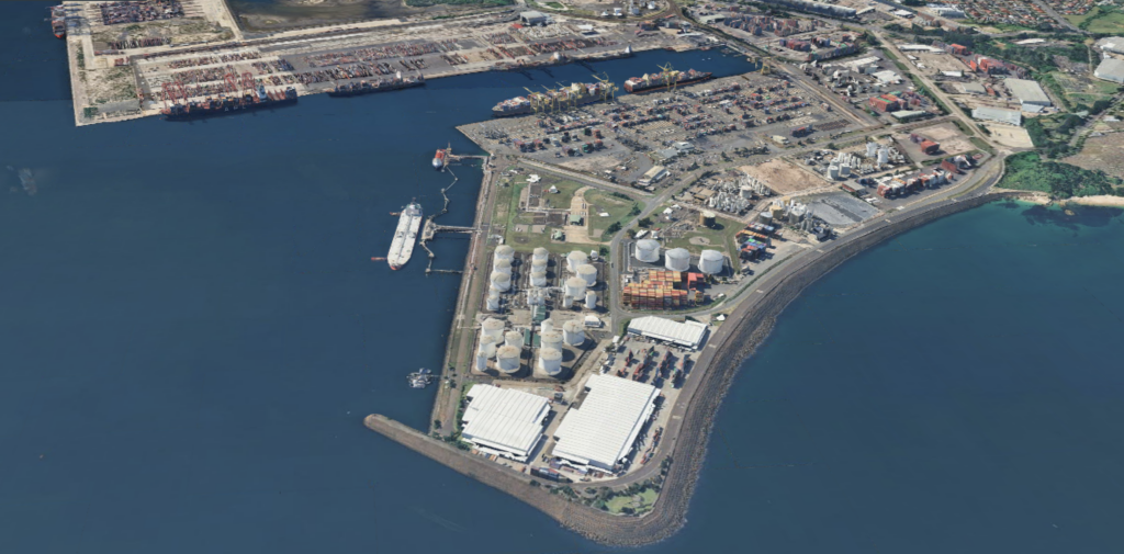 Aerial view of Sydney's industrial port with storage tanks, warehouses, shipping containers, and a docked cargo ship along the coastline.