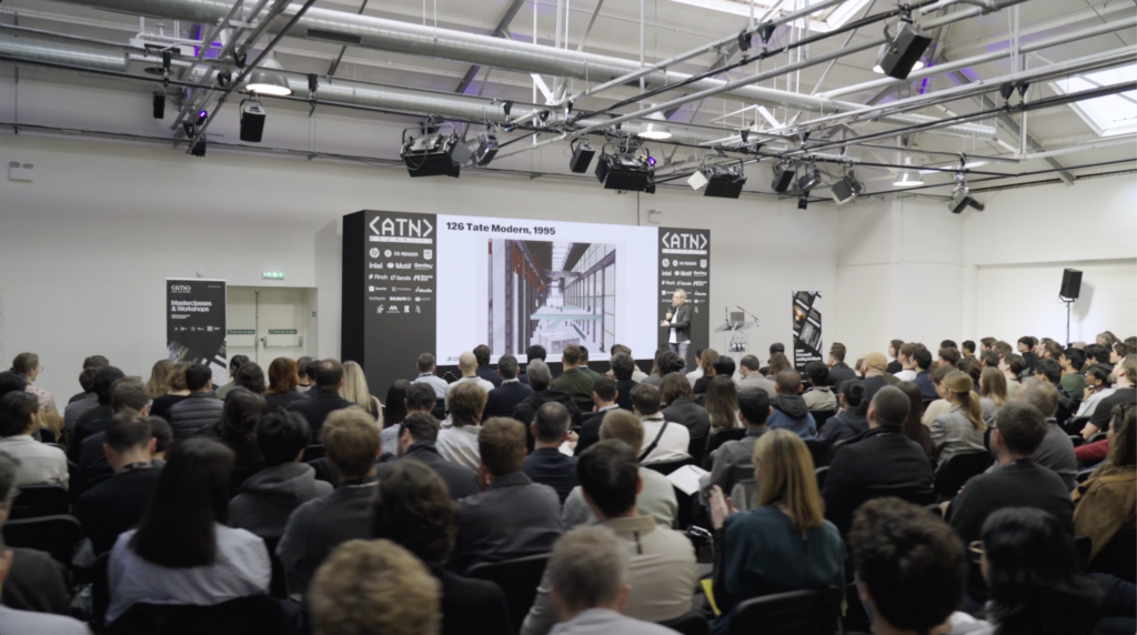 A large audience sits facing a presenter, likely an architect, who stands next to a screen displaying an architectural image with the title Tate Modern, 1995 in a bright conference room.