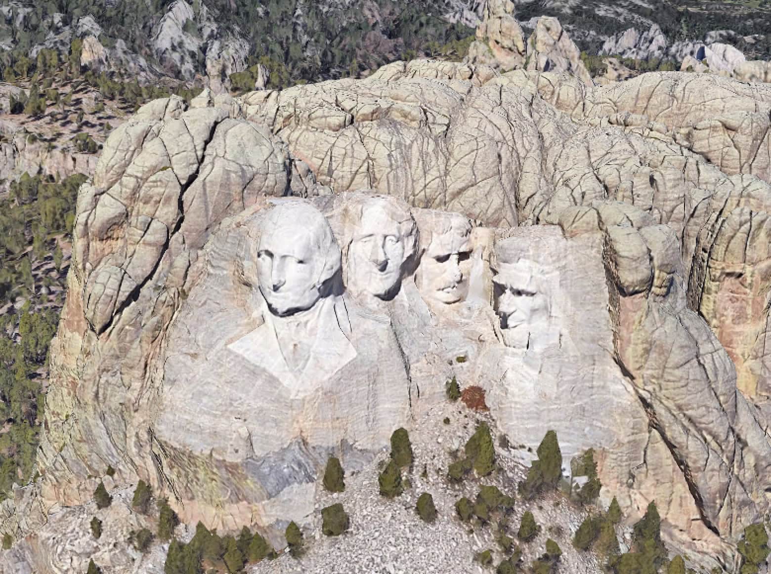 Aerial view of Mount Rushmore showing four large carved presidential faces in a granite mountain, surrounded by trees and rocky terrain.