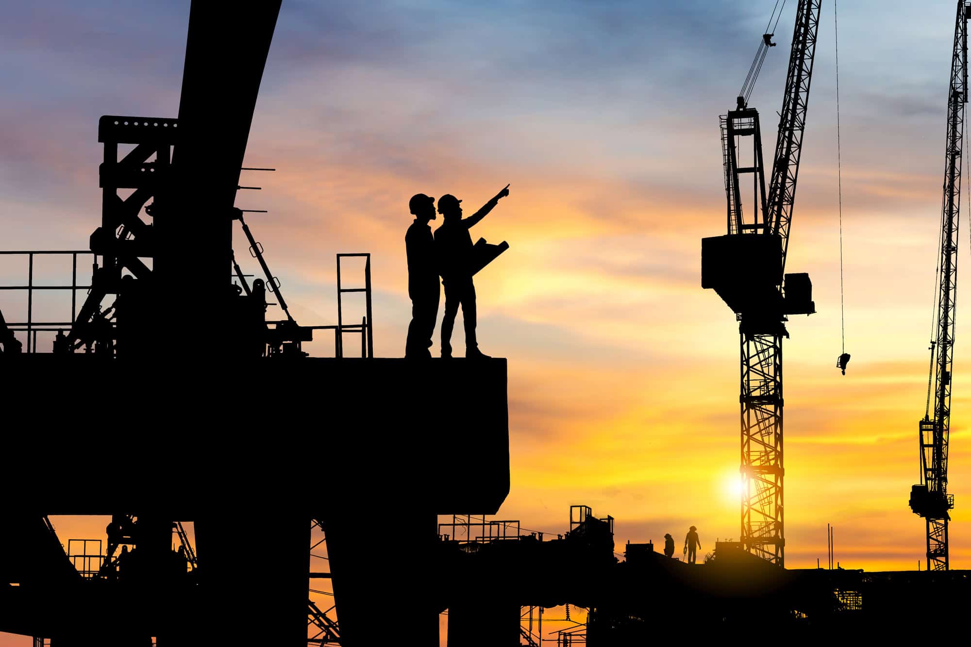Silhouetted construction workers on a site at sunset, with cranes and building structures in the background. One worker points towards the horizon while discussing plans.