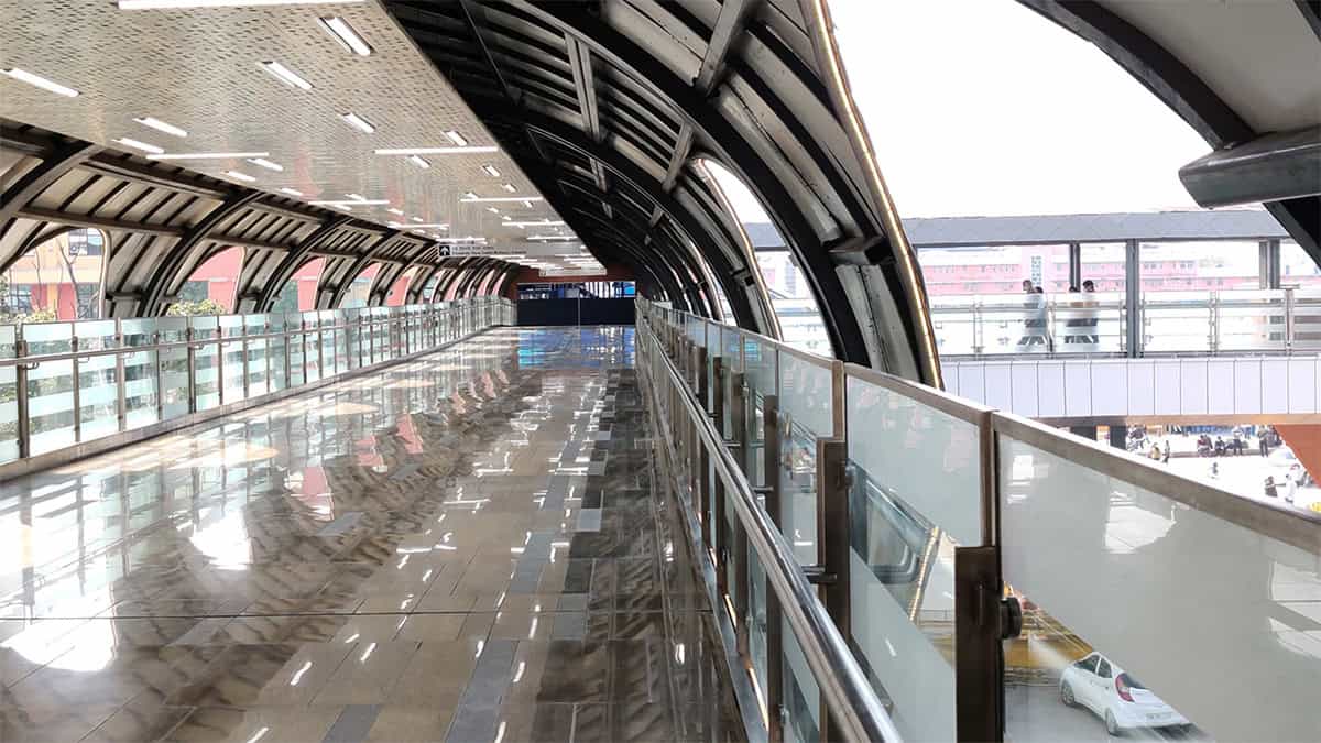 A modern, covered pedestrian walkway with glass railings and a reflective floor, designed by AVS ENGINEERS, extends toward a bright outdoor area at Fairmont Palace Udaipur, with another walkway visible in the distance.