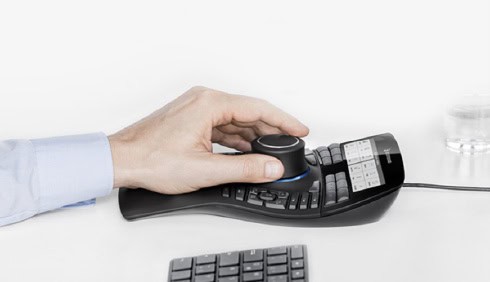 A person's hand operates a 3D mouse with multiple buttons, next to a standard keyboard on a white desk&mdash;perfect for CAD tasks in Infrastructure Engineering or working with MicroStation 2026.