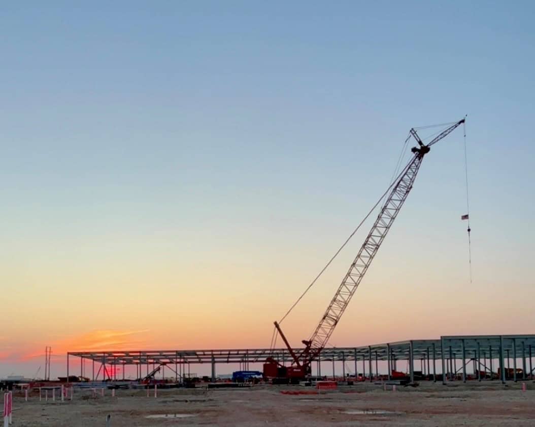 A tall crane stands at a Texas construction site, where DPR Construction is building steel framework structures for hyperscale AI data centers at sunset under a clear sky.
