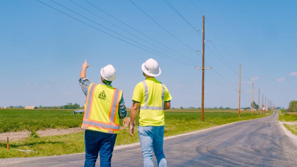 Two engineers in safety vests and hard hats stand on a rural road, pointing at overhead power lines that run alongside fields under a clear sky.