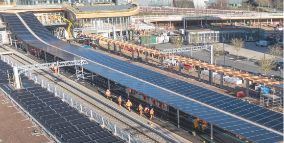 A railway station with multiple train tracks covered by large solar panels, featuring the Arcadis Rail Division&rsquo;s commitment to engineering clarity as several workers in orange vests walk along the tracks.