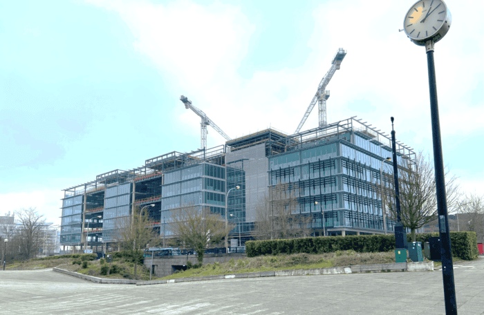 A large modern office building under construction with two cranes on top. A streetlamp with a clock is in the foreground and trees are around the building.