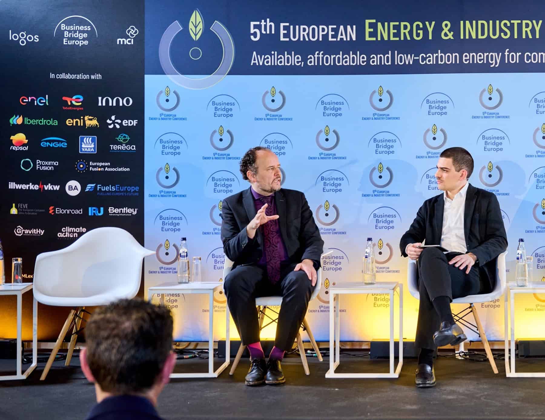 Two men in business attire sit on stage discussing at the 5th European Energy and Industry event.